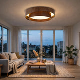 A modern circular ceiling light fixture with a natural wood outer ring and a glowing white inner diffuser, installed in a bright, contemporary living room with large windows and a sofa.