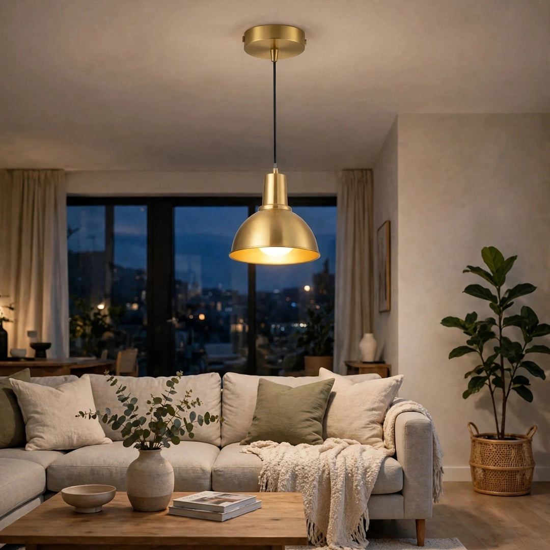 A modern brass pendant light with a conical shade hanging over a cozy living room with a beige sofa, wooden coffee table, and a large potted plant.