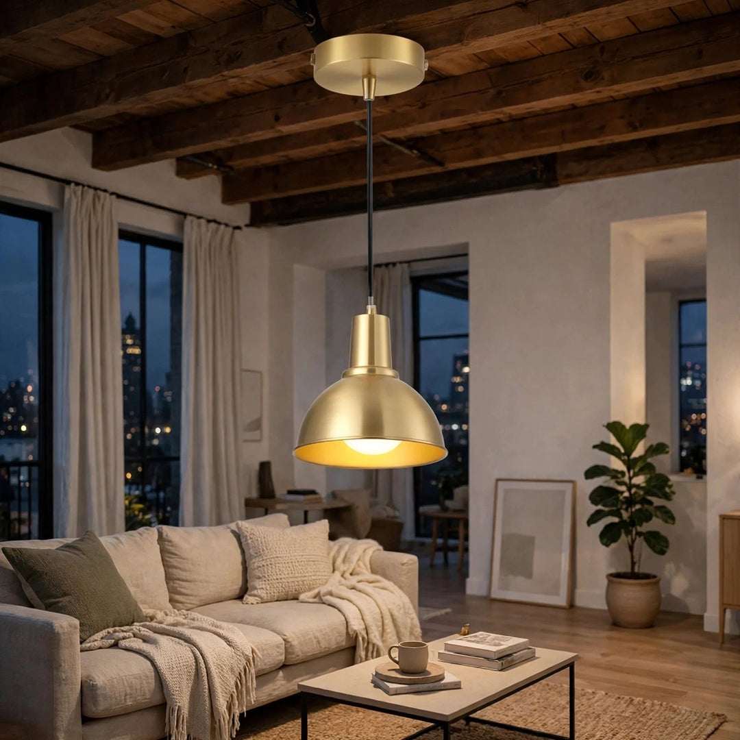 A modern pendant light with a brushed gold dome shade and a black cord hanging in a stylishly decorated living room with wooden ceiling beams, a beige sofa, and large windows overlooking a city skyline at dusk.