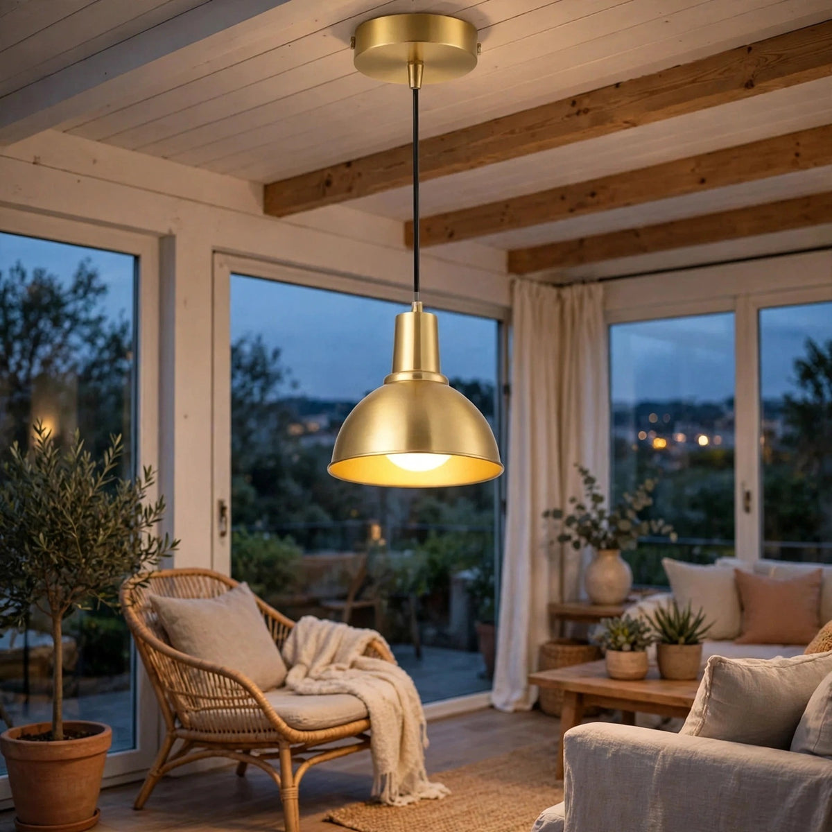 A modern pendant light with a brushed gold dome shade and black cord hanging in a cozy, sunlit living room with wood ceiling beams, a wicker chair, and large windows overlooking a garden at dusk.