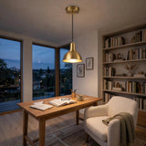 A modern brass dome pendant light hangs over a wooden desk in a cozy home office with a white armchair, bookshelves, and large windows showing dusk outside.