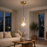 A modern pendant light with a brushed gold brass cylinder and a clear glass globe bulb, hanging in a cozy, warmly lit living room with a sofa and wooden coffee table.