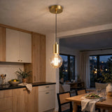 A modern pendant light with a brushed gold ceiling canopy and socket, a black cord, and a clear glass globe bulb glowing with warm light, hanging in a contemporary kitchen and dining room.