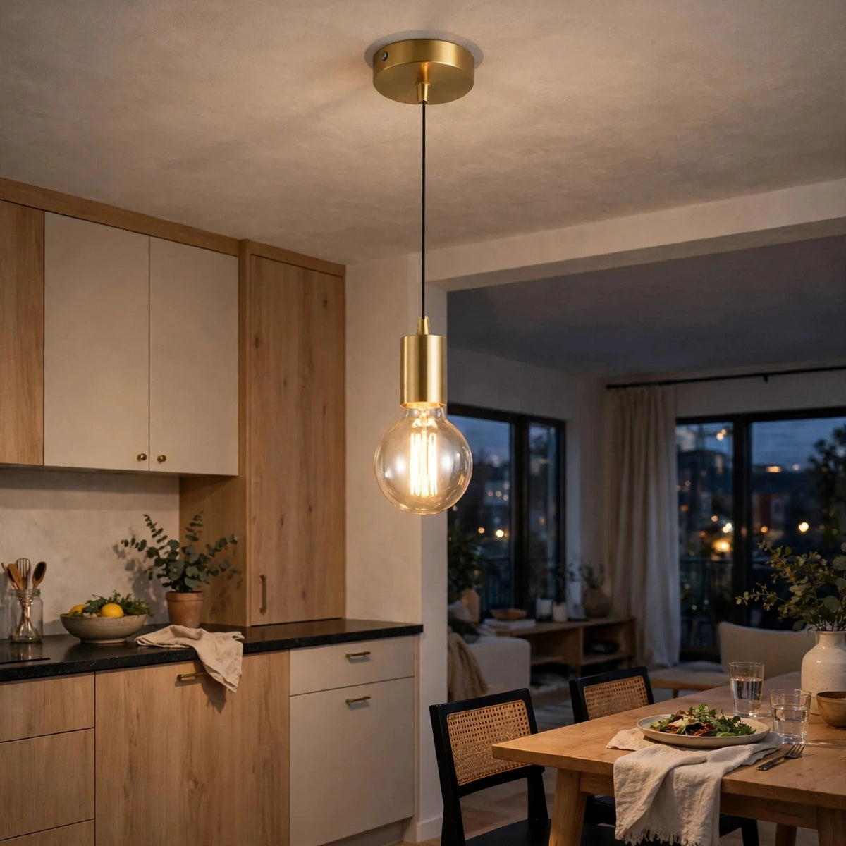 A modern pendant light with a brushed gold ceiling canopy and socket, a black cord, and a clear glass globe bulb glowing with warm light, hanging in a contemporary kitchen and dining room.