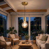 A modern pendant light with a brushed brass cylindrical collar and a clear glass globe bulb hanging in a cozy, sunlit conservatory with wicker chairs, a sofa, and wooden ceiling beams.