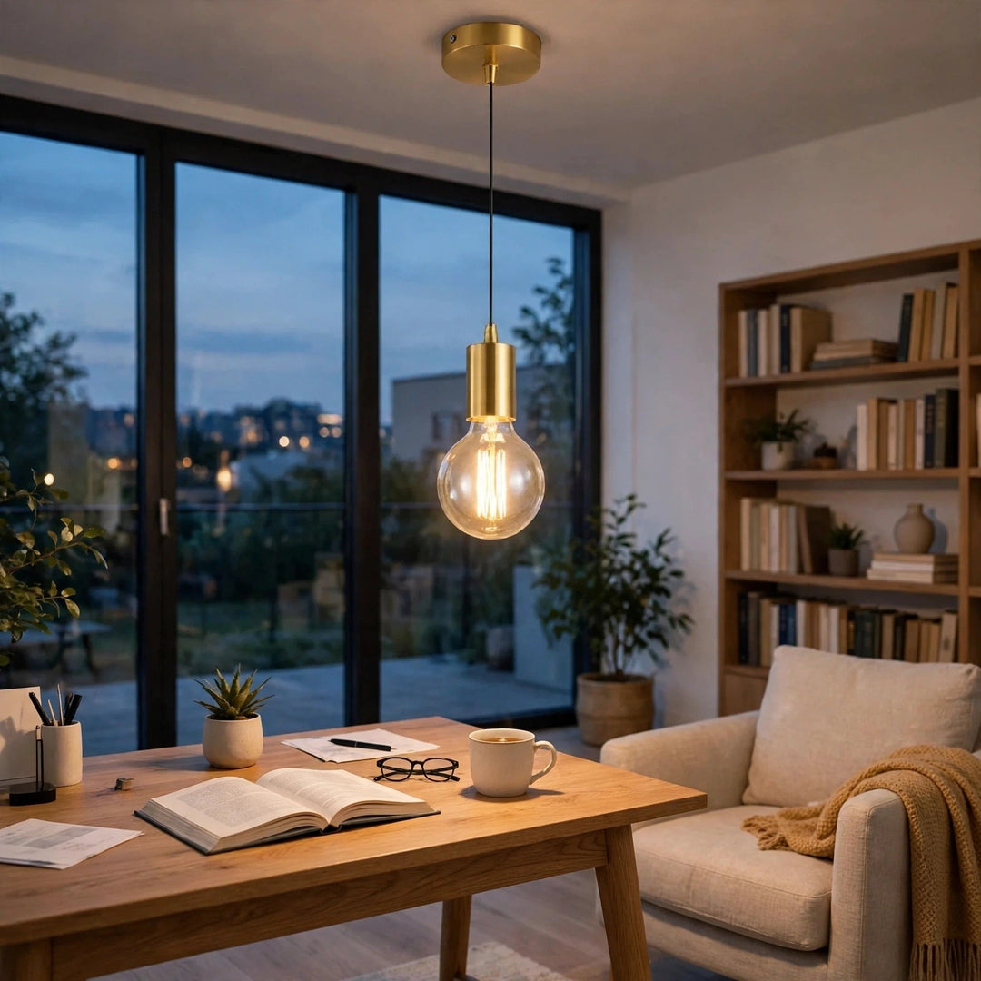 A modern brass pendant light with a clear glass globe bulb hanging over a wooden desk in a cozy, well-lit home office.