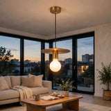 A modern pendant light with a wide brass-colored metal disc shade and a glowing warm white globe bulb, hanging in a bright living room with large windows overlooking a city skyline at dusk.