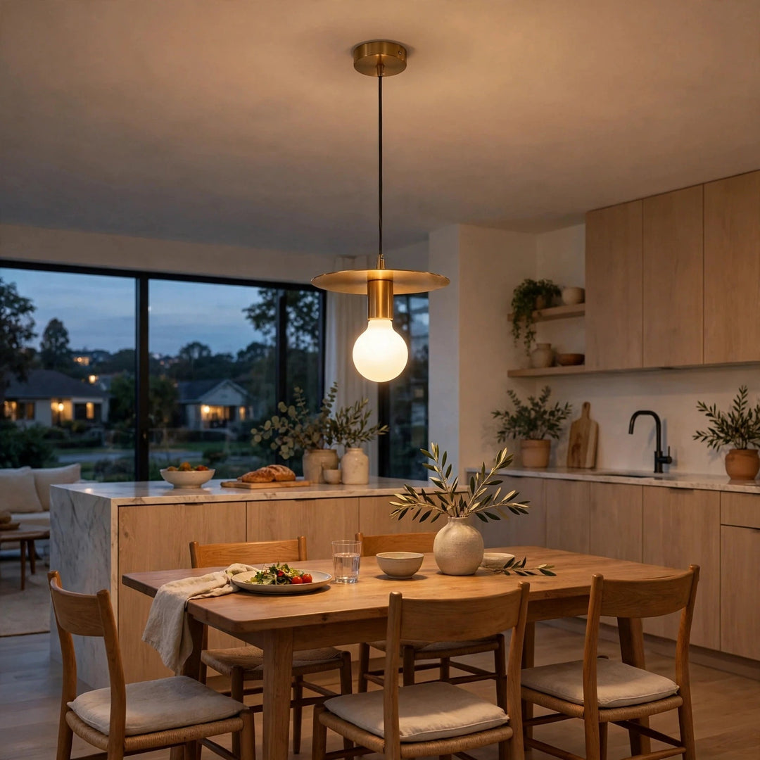 A modern brass pendant light with a frosted glass globe bulb hangs over a wooden dining table in a contemporary kitchen with light wood cabinets and large windows showing an evening outdoor view.