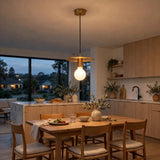 A modern brass pendant light with a frosted glass globe bulb hangs over a wooden dining table in a contemporary kitchen with light wood cabinets and large windows showing an evening outdoor view.