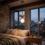 A modern pendant light with a brass-toned metal shade and a glowing warm white globe bulb hangs over a bed in a styled bedroom with an exposed brick wall, large windows, and a city view at dusk.