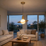 A modern pendant light with a brushed brass finish and a glowing frosted globe shade, suspended in a cozy, contemporary living room with beige sofas and large windows.