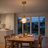 A modern brass pendant light with a glowing frosted globe shade hangs over a wooden dining table in a bright, contemporary kitchen and dining room.