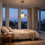 A modern pendant light with a wide circular shade and a glowing globe bulb hangs over a neatly made bed in a softly lit bedroom with large windows and city views at dusk.