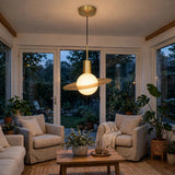 A modern pendant light with a glowing white globe and a gold-toned ring shade hangs in a cozy, sunlit living room with two armchairs, a wooden coffee table, and large windows overlooking a garden.