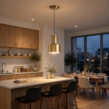 A modern kitchen and dining room interior with a brushed gold pendant light hanging above a kitchen island. The space features light wood cabinetry, a marble countertop, black bar stools, and a dining table with chairs near large floor-to-ceiling windows showing an evening city view.