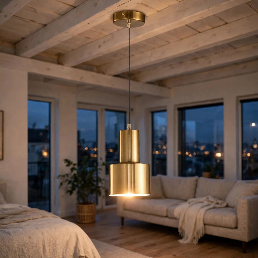 A modern two-tiered pendant light in brushed gold metal hangs in a cozy, contemporary loft bedroom with exposed wood ceiling beams, large windows, and a light beige sofa.