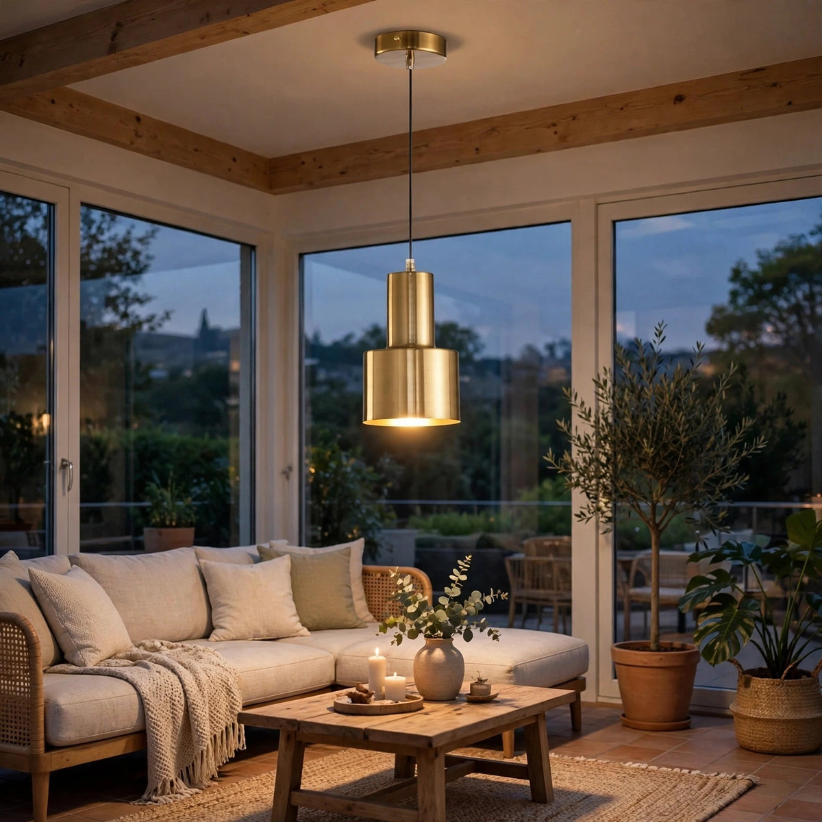 A modern two-tiered brass pendant light hangs in a bright, sunlit living room with a beige sofa, wooden ceiling beams, and large windows overlooking a garden at dusk.