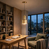 A modern brass pendant light hangs over a wooden desk in a cozy home office with a bookshelf, armchair, and large windows overlooking a city at dusk.