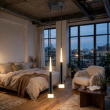 Two modern pendant lights with slim black conical shades and glowing white LED ends hang over a bed in a styled loft bedroom with large windows, a bed, and a chair.