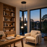 A modern three-light linear pendant chandelier with glowing cylindrical bulbs hangs over a wooden desk in a cozy home office. The room features a large wooden bookshelf, a cream-colored armchair with a throw blanket, and large floor-to-ceiling windows showing a twilight city view.