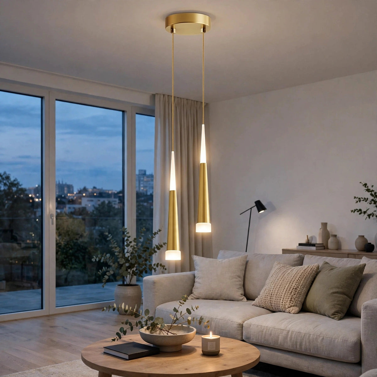 A modern living room with a three-light linear pendant ceiling fixture in brushed gold and white, hanging above a light beige sofa. Large windows in the background show a twilight city view.