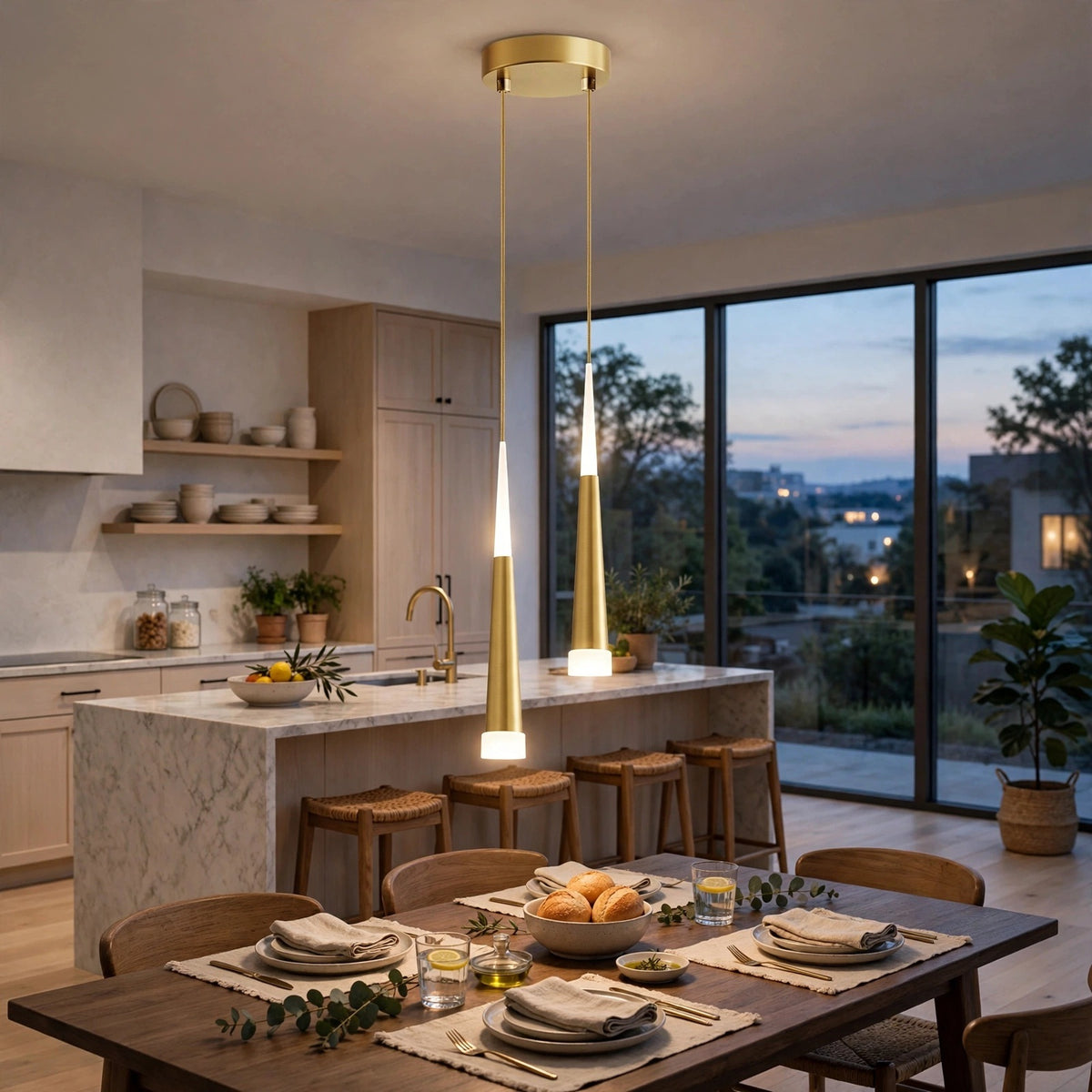 A modern kitchen and dining room interior with a two-light pendant fixture hanging over a dining table. The fixture has a gold-toned circular canopy and two slim tapered shades that glow warm white when lit.