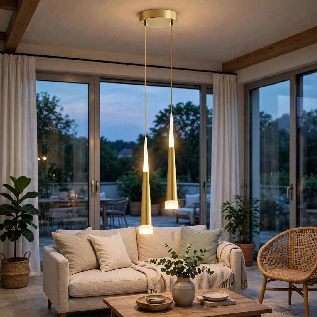 A modern two-light pendant ceiling fixture with brushed gold/brass cylindrical shades and warm white glowing bulbs, hanging in a styled living room with a sofa, plants, and large windows.