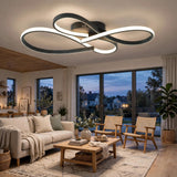 Modern ceiling light fixture with an infinity-shaped black frame and glowing white LED strips, installed in a contemporary living room with a sofa, coffee table, and large windows.