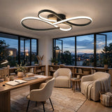 A modern infinity-shaped LED ceiling light with a black frame and glowing white light, installed in a stylish contemporary home office with large windows, bookshelves, and beige armchairs.