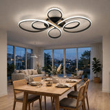 Modern ceiling light fixture with multiple interlocking circular rings in a black and brushed metal finish, illuminated in a contemporary dining room with a wooden table, chairs, and large windows overlooking a city view at dusk.