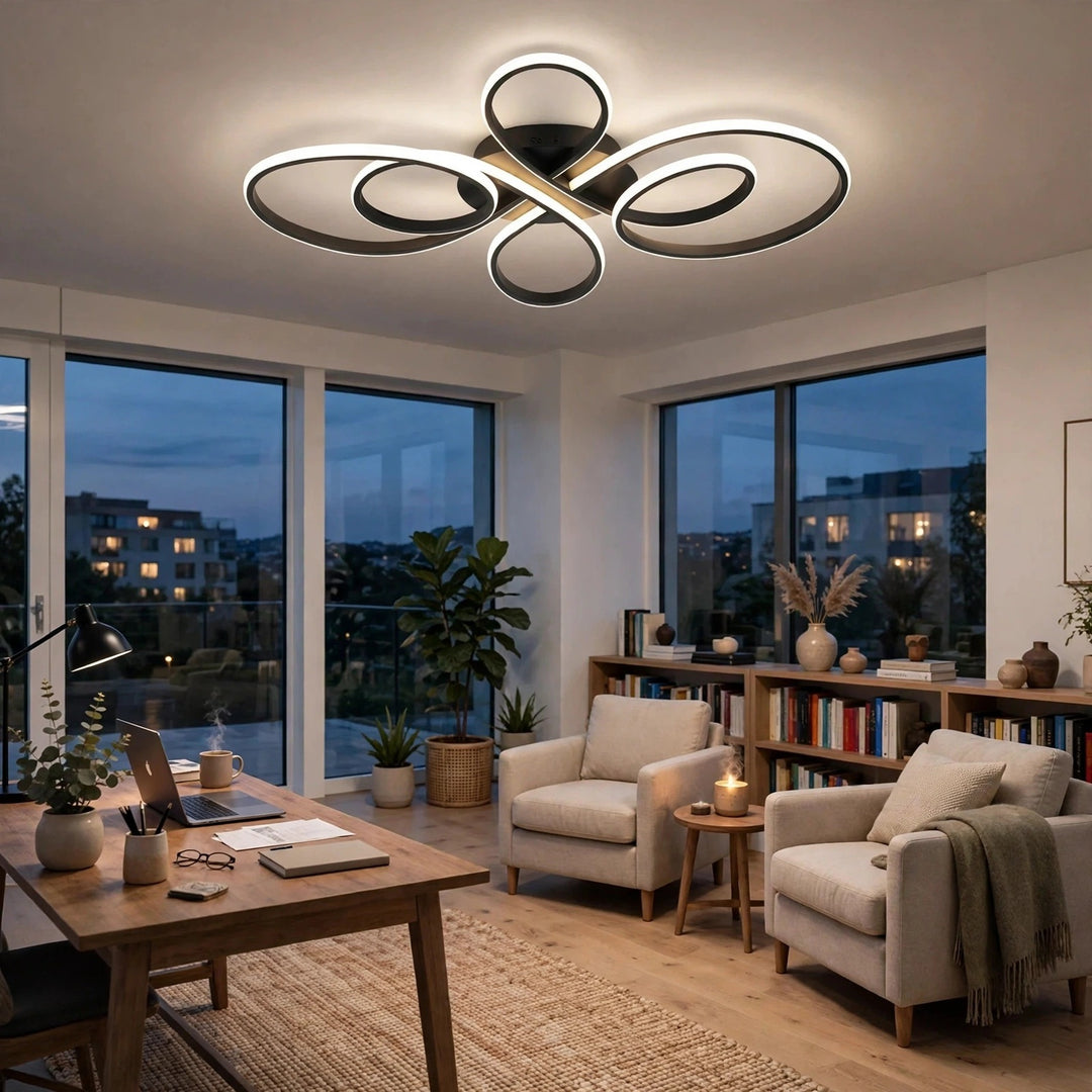 Modern ceiling light fixture with interlocking circular rings in black and brushed silver, glowing with warm white light in a contemporary living room with beige armchairs, a wooden desk, and large windows.