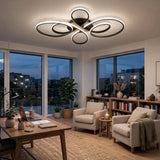 Modern ceiling light fixture with interlocking circular rings in black and brushed silver, glowing with warm white light in a contemporary living room with beige armchairs, a wooden desk, and large windows.