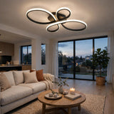 Modern ceiling light fixture with interlocking oval rings in black and brushed metal, glowing with warm white light in a contemporary living room with a sofa, coffee table, and large windows.