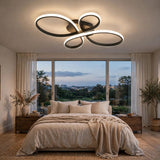 Modern ceiling light fixture with three interlocking oval rings in a black finish, emitting a warm white glow, installed in a contemporary bedroom with a bed, large windows, and plants.