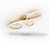 A gold ceiling light is viewed at an angle, showing curling illuminated bands and its raised central mount against white.