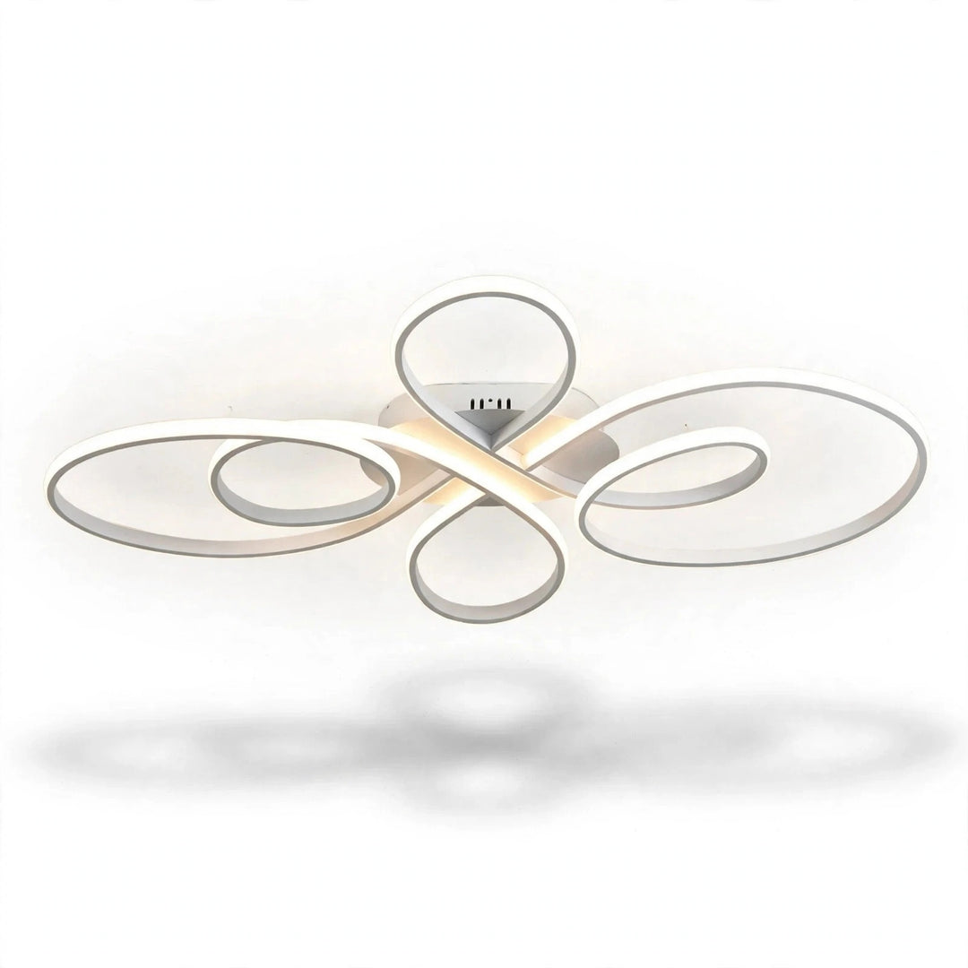 A modern ceiling light fixture with flowing, abstract oval loops in a brushed silver finish, emitting a warm white glow from its central LED light source, shown against a plain white background with a soft shadow below.