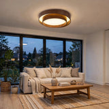 A modern circular ceiling light with a dark wood outer ring and a glowing white inner ring, installed in a bright living room above a beige sofa and wooden coffee table.