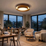 A modern circular ceiling light with a warm wood frame and glowing white diffuser, installed in a stylishly decorated living room with beige armchairs, a wooden desk, and large windows overlooking a city at dusk.