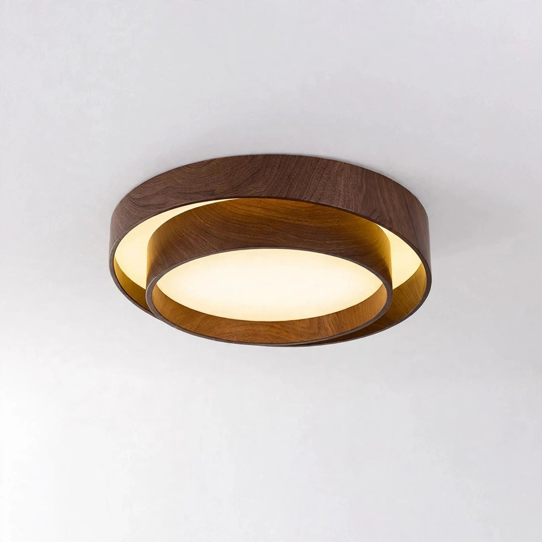 A modern flush-mount ceiling light fixture with concentric circular rings in dark brown wood and a warm white illuminated diffuser, shown against a plain light gray ceiling.