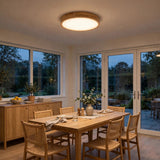 A modern flush-mount ceiling light with a circular wooden frame and warm white glow, installed in a bright, contemporary dining room with light wood furniture and large windows overlooking a garden.