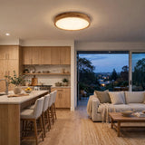 A modern circular ceiling light with a natural wood rim and warm glowing light, installed in a contemporary open-plan kitchen and living room.