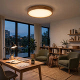 A modern circular flush-mount ceiling light with a natural wood rim and warm white glow, installed in a cozy, contemporary home office with green armchairs, a wooden desk, and large windows overlooking a city skyline at dusk.