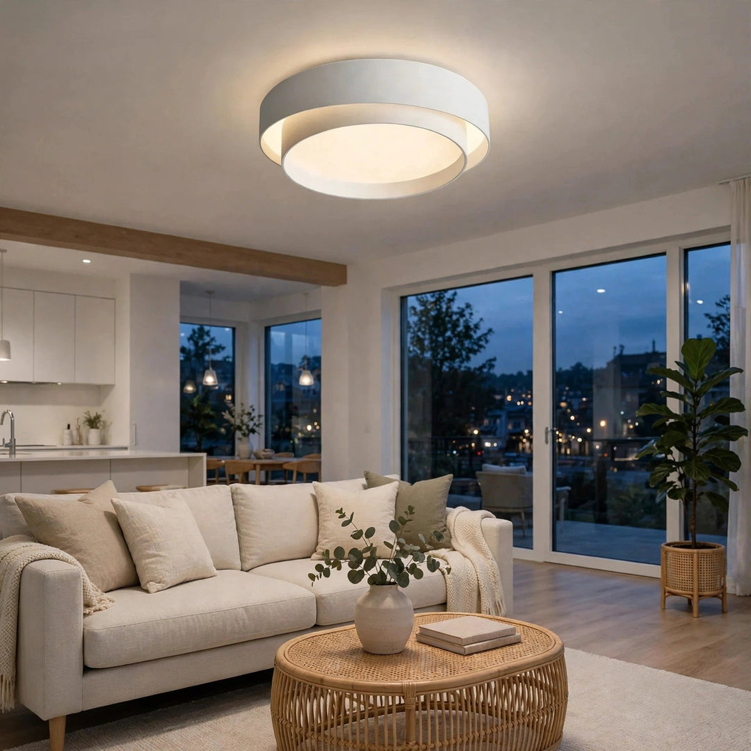 A modern circular flush-mount ceiling light with a white outer ring and a glowing warm white inner ring, installed in a bright, contemporary living room with a beige sofa and large windows.