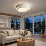 A modern circular flush-mount ceiling light with a white outer ring and a glowing warm white inner ring, installed in a bright, contemporary living room with a beige sofa and large windows.