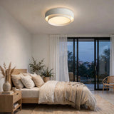 A modern circular flush-mount ceiling light with a white outer ring and a warm glowing white diffuser, installed in a minimalist, warm-toned bedroom.