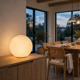 White glass globe wall sconce displayed on a wooden sideboard in a dining room with woven chairs and large windows.