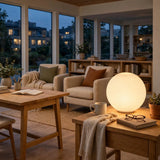 White glass globe wall sconce displayed on a wooden table beside a mug and books, with beige armchairs behind.