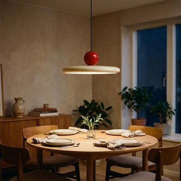 A modern pendant light with a circular beige shade and a glossy red spherical bulb, hanging over a wooden dining table set for four in a warmly lit dining room.