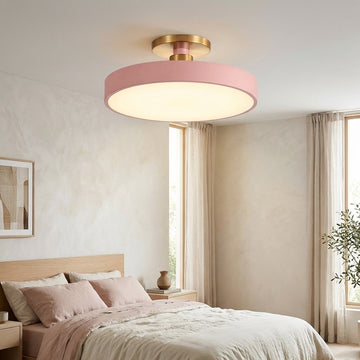 A modern semi-flush mount ceiling light with a pink cylindrical shade and gold-toned metal hardware, illuminated in a softly lit bedroom setting.