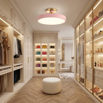 A modern walk-in closet with cream cabinetry, gold-framed glass doors, built-in shelving with colorful handbags, a round white ottoman, and a glowing circular ceiling light.